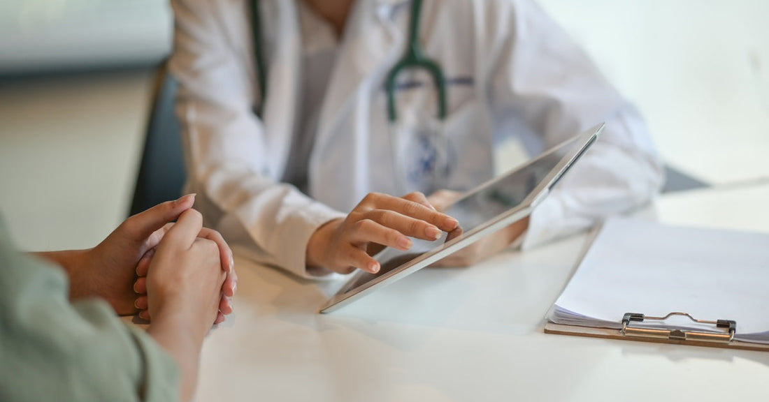 A doctor sitting at a table across from their patient. The doctor is holding and tapping a white tablet.