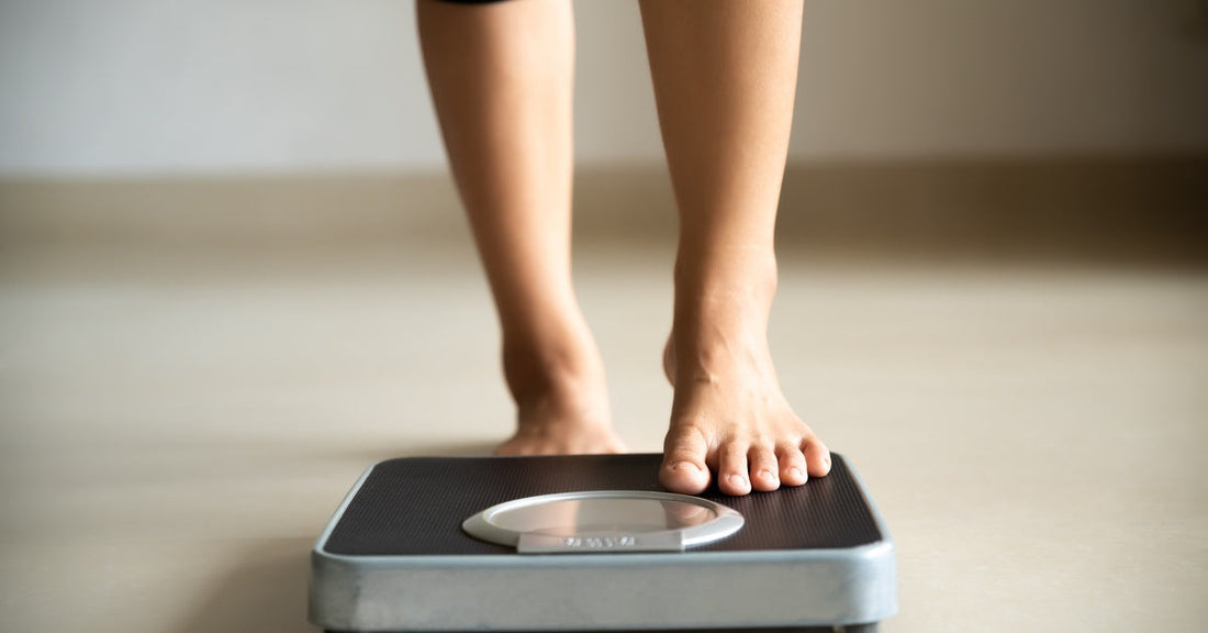 A person wearing black pants placing their left foot on a small black-and-gray scale. The scale is on the floor.