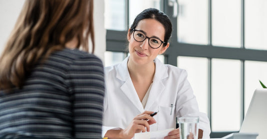 A doctor sitting at a desk in their office with a patient. The doctor smiles and holds a pen in their right hand.