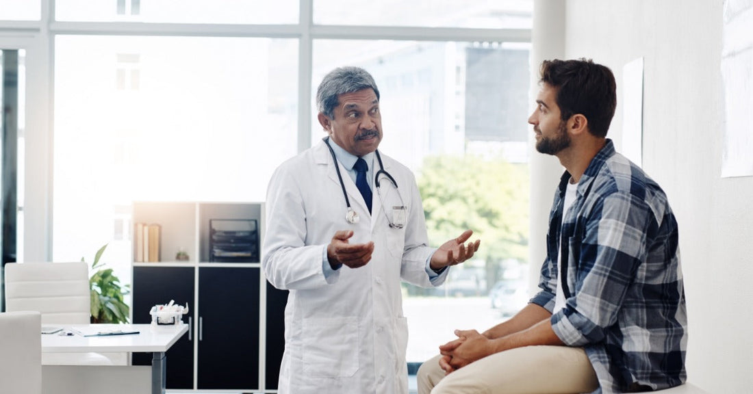 A doctor standing next to a patient sitting on a bench in their office. Large windows let the sun inside the room.