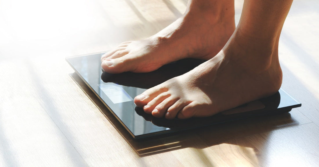 A person standing with their bare feet on a small back scale on the floor. Sunlight casts shadows over the scale.
