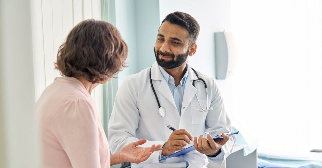 A patient wearing a pink shirt talking to a doctor in their office. The doctor holds a pen and clipboard in their hands.