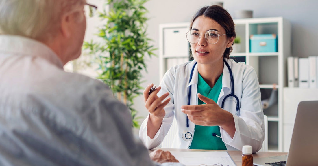 A doctor wearing glasses sitting at a desk in their office. The doctor talks to a patient sitting across from them.