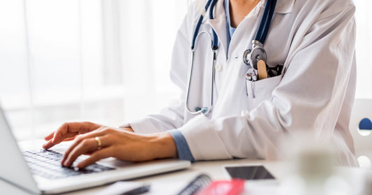A doctor sitting at a desk in their office. The doctor types on a small white laptop sitting on the desktop.