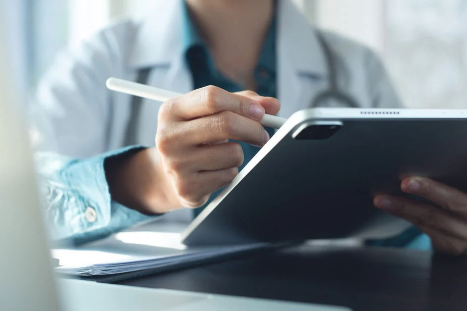 A doctor sitting in their office during the daytime and writing with a white stylus on a small gray tablet