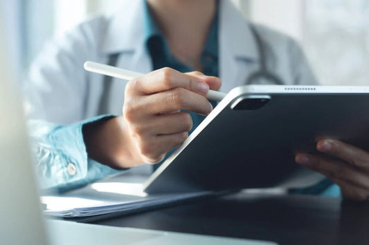 A doctor sitting in their office during the daytime and writing with a white stylus on a small gray tablet